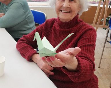Ogród zimowy. Warsztaty origami. Seniorka prezentuje ptaka złożonego z papieru.