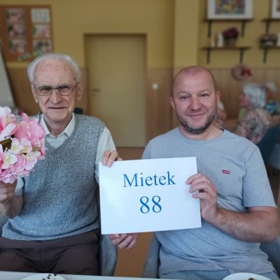 Sala. Na pierwszym planie kierownik Arkadiusz Wanat i senior. Trzymają kartkę z napisem Mietek 88. Senior ma bukiet kwiatów.