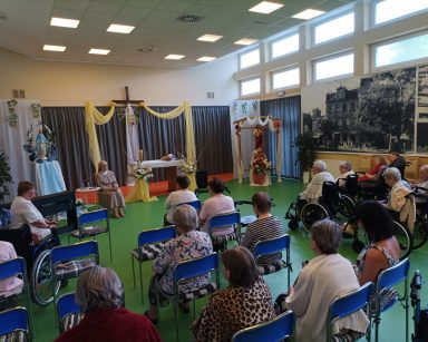 Sala. Siostra Maria Luiza prowadzi modlitwy. Przed nią siedzą seniorzy. Z boku siedzi wolontariuszka pani Jagoda.