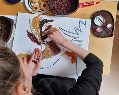 Widok z góry. Dziewczyna wykleja obraz koguta z kolorowych ziaren i nasion.