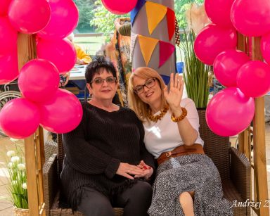 Ogród zimowy. Pergola z różowymi balonami. Na kanapie siedzi seniorka i dyrektor Agnieszka Cysewska. Uśmiechają się.