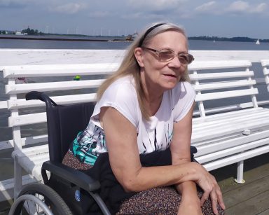 Molo w Pucku. Seniorka siedzi na wózku inwalidzkim. Za nią widać morze i linię brzegową.