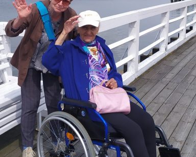Pogodny dzień. Molo w Pucku. Wolontariuszka i seniorka na wózku inwalidzkim spacerują po molo.