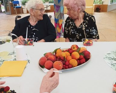 Ogród zimowy. Przy stole siedzą seniorzy. Jedzą świeże owoce, rozmawiają.