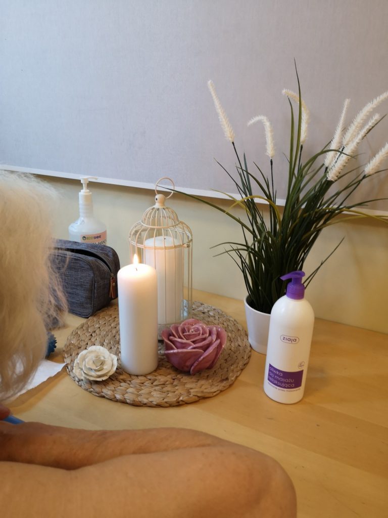 Kobieta opiera się o stół. Na blacie dekoracja, świece, wazon z bukietem, oliwka do masażu.
