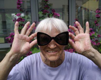 Ogród. Seniorka mierzy ciemne okulary. Śmieje się.