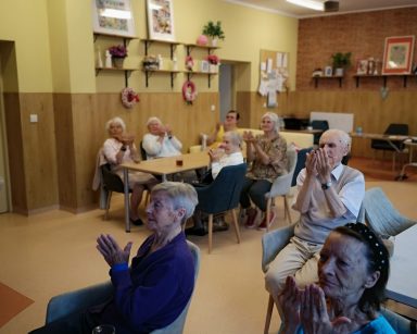 Sala. Seniorzy siedzą przy stołach. Unoszą ręce, wykonują ćwiczenia dłoni.