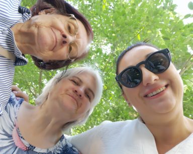Do zdjęcia pozuje terapeutka Gosia Jancelewicz i dwie seniorki. Uśmiechają się. W tle drzewo z zielonymi liśćmi.
