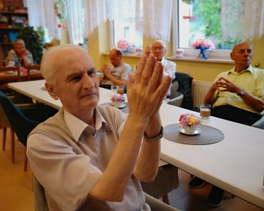 Sala. Seniorzy siedzą przy stołach. Unoszą ręce, wykonują ćwiczenia dłoni.