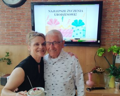 Sala. Terapeutka Beata Brzozowska i senior stoją objęci. Uśmiechają się. Za nimi ekran i napis Najlepsze życzenia urodzinowe.