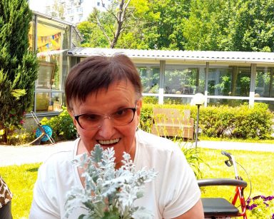 Ogród. Słoneczny dzień. Seniorka trzyma w rękach kwiat mrozy. Śmieje się.