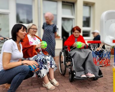 Patio przed budynkiem. Terapeutka Ani Rzepczyńska i seniorki trzymają kolorowe piłki. Przed nimi mały kosz do koszykówki.