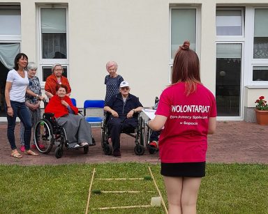 Terapeutka Ania Rzepczyńska i seniorzy na patio przed budynkiem. Na trawniku stoi wolontariuszka.