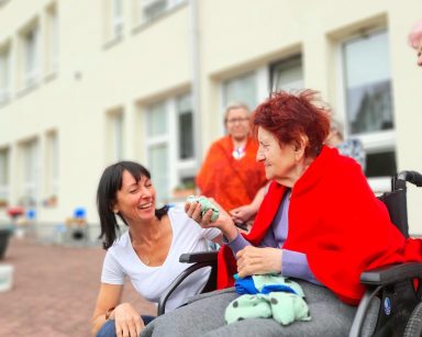 Patio przed budynkiem. Na pierwszym planie terapeutka Ania Rzepczyńska. Obok seniorka na wózku inwalidzkim.