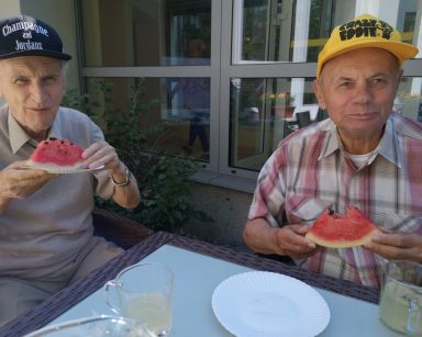 Na świeżym powietrzu przy stole siedzą dwaj seniorzy. Jedzą arbuza.