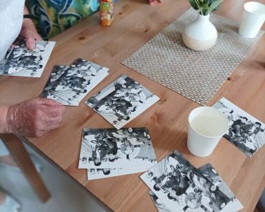Sala. Na blacie stołu rozłożone biało-czarne fotografie. Seniorzy biorą je do rąk, oglądają.