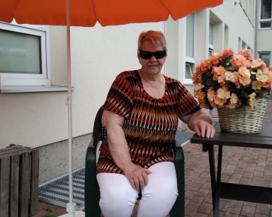 Patio przed budynkiem. Seniorka siedzi na krześle, pod parasolem. Ma ciemne okulary.