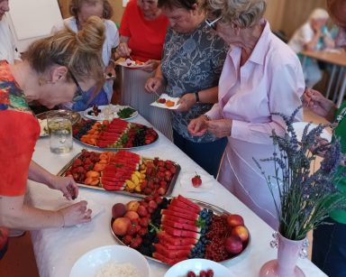 Sala. Na stole tace z owocami. Seniorzy nakładają sobie owoce na tacki.