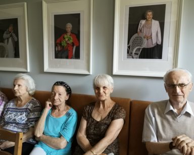 Sala. Seniorzy siedzą na kanapie pod ścianą. Słuchają. Na ścianie wiszą portrety oprawione w ramki.
