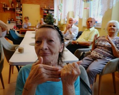 Sala. Seniorzy siedzą przy stołach. Unoszą ręce, wykonują ćwiczenia dłoni i palców.