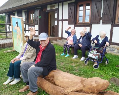 Słoneczny dzień. Trawnik przed białą chatą. Na drewnianych ławach siedzą seniorzy. Machają do zdjęcia.