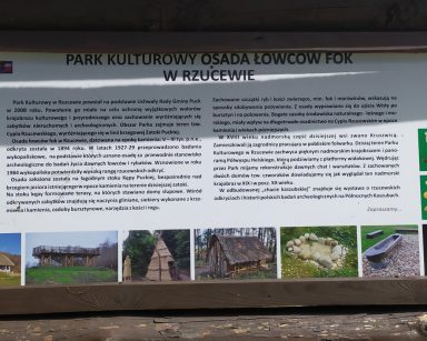 Tablica w drewnianej ramie na niej tekst Park Kulturowy Osada Łowców Fok w Rzucewie. Pod napisem opis i zdjęcia.