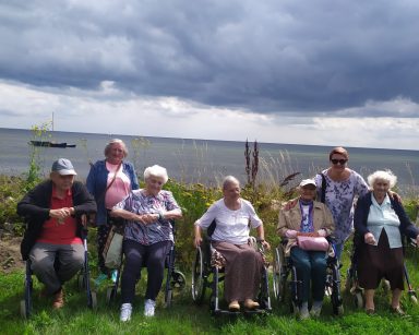Grupa seniorów i koordynatorka Edyta Życzyńska pozują do zdjęcia na świeżym powietrzu. W tle łąka z kwiatami, dalej morze.