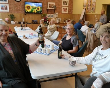 Sala. Przy stolach siedzą seniorzy. Śmieją się, rozmawiają. Jedna z seniorek podnosi butelkę z kwasem chlebowym.