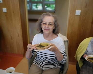 Sala. Seniorka siedzi przy stole. Uśmiecha się. Trzyma talerzyk z kanapką ze smalcem i ogórkiem.