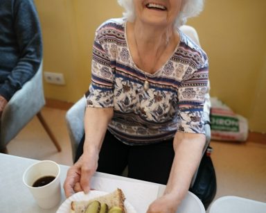 Sala. Seniorka siedzi przy stole. Śmieje się. Przed nią talerzyk z kanapką ze smalcem i ogórkiem, kubek z napojem.