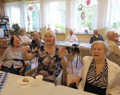 Sala. Seniorzy siedzą przy stołach. Jedzą, śmieją się, rozmawiają.