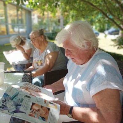 Słoneczny dzień. Ogród. W cieniu drzew siedzą seniorki. Czytają prasę.