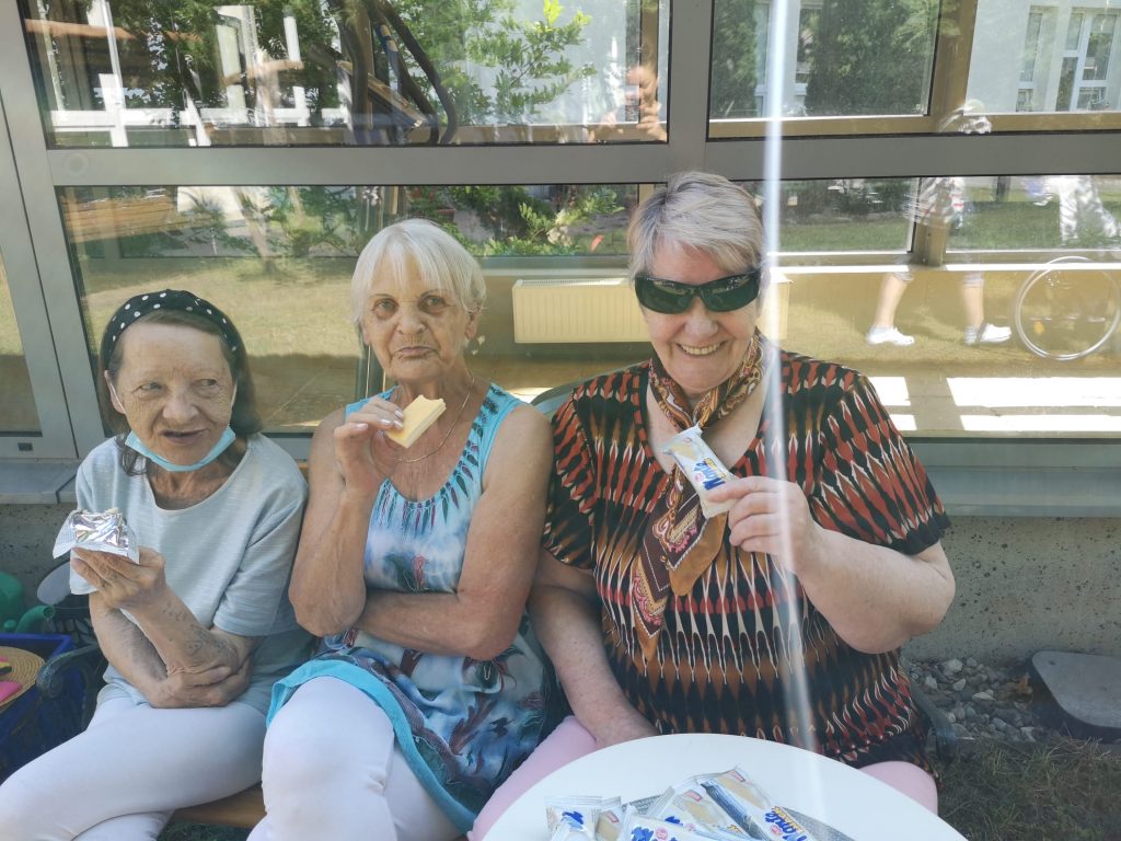 Słoneczny dzień. Na świeżym powietrzy powietrzu trzy seniorki. Siedzą na ławce, jedzą słodycze.