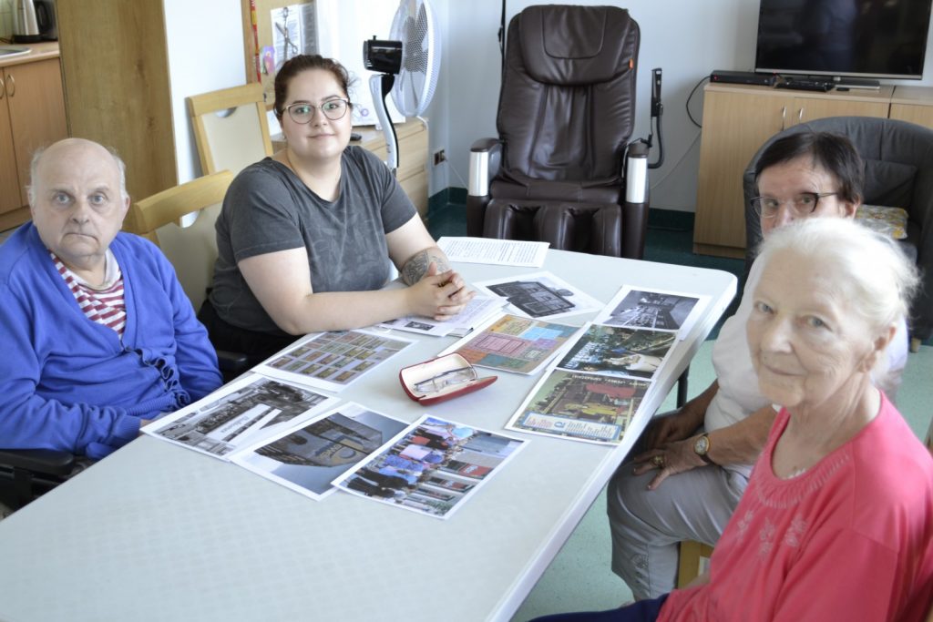 Pokój. Psycholożka Bogumiła Syposz prowadzi zajęcia z seniorami. Siedzą przy stole, na blacie zdjęcia.