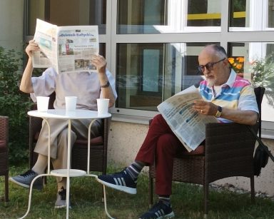 Słoneczny dzień. Na świeżym powietrzu, przy stoliku siedzą dwaj seniorzy. Czytają prasę.