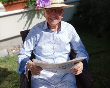 Słoneczny dzień. Na świeżym powietrzu, na rattanowym fotelu siedzi senior. Śmieje się, przegląda gazetę.