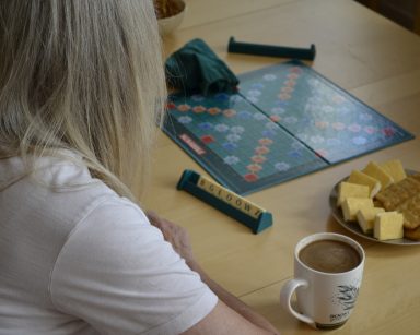Świetlica. Seniorka siedzi przy stole. Przed nią plansza do gry w scrabble, ciastka i kawa.