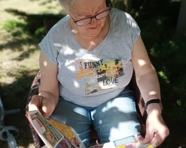Słoneczny dzień. Na świeżym powietrzu, w cieniu drzewa siedzi seniorka. Czyta gazetę.