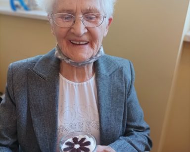 Sala. Seniorka siedzi na krześle. Śmieje się. Trzyma kubek z kawą.