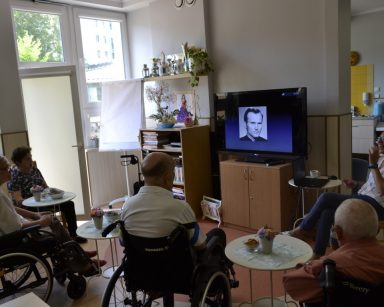 Świetlica. Terapeutka Magdalena Poraj-Górska prowadzi spotkanie. Przed nią siedzą seniorzy. Patrzą na zdjęcie Jana Pawła II.