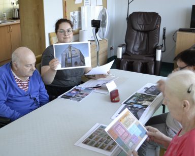 Pokój. Psycholożka Bogumiła Syposz prowadzi zajęcia z seniorami. Siedzą przy stole, na blacie zdjęcia.