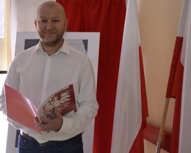 Kierownik Arkadiusz Wanat stoi ze śpiewnikiem. Za nim dekoracja z okazji rocznicy powstania. Flagi Polski.