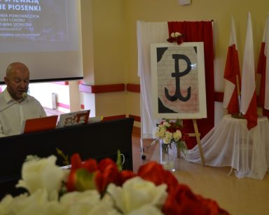 Kierownik Arkadiusz Wanat gra na pianinie. Obok dekoracja w kolorze biało-czerwonym, kwiaty, flagi, symbol Polski walczącej.