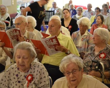 Seniorzy i pracownicy siedzą w rzędach. Mają biało-czerwone opaski na ramię, kotyliony i śpiewniki z orłem.