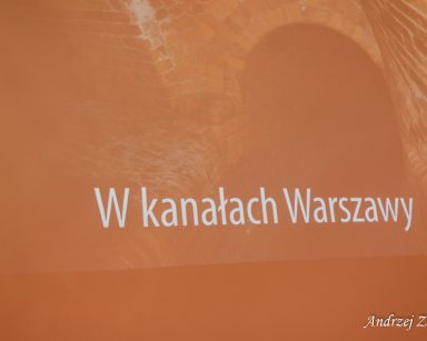 Ekran projektora. Obraz ściany z cegieł, wejście do kanału, niżej napis W kanałach Warszawy.