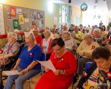 Seniorzy i pracownicy siedzą w rzędach. Mają biało-czerwone opaski na ramię, kotyliony i śpiewniki z orłem.