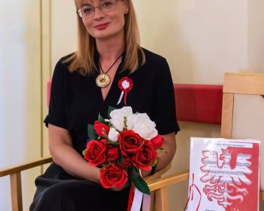 Dyrektorka Agnieszka Cysewska siedzi na krześle. Uśmiecha się. Trzyma bukiet białych i czerwonych róż. Obok leży śpiewnik.