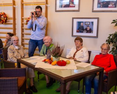 Sala. Przy stołach siedzą seniorzy i wolontariuszki. Administrator Łukasz Cielebański robi zdjęcia.