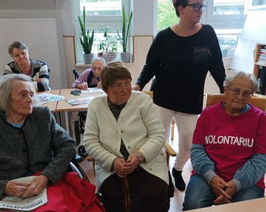 Świetlica. Przy stołach siedzą seniorzy, wolontariusze. Za krzesłami stoi koordynatorka Edyta Życzyńska.