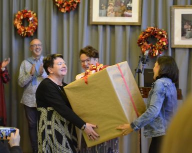 Sala. Trzy osoby trzymają ogromny prezent. Prezent jest zapakowany w brązowy papier i przewiązany wstążką.